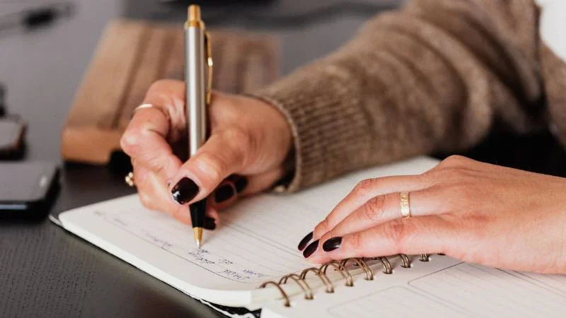 Person writing in a notebook with a pen on a desk.
