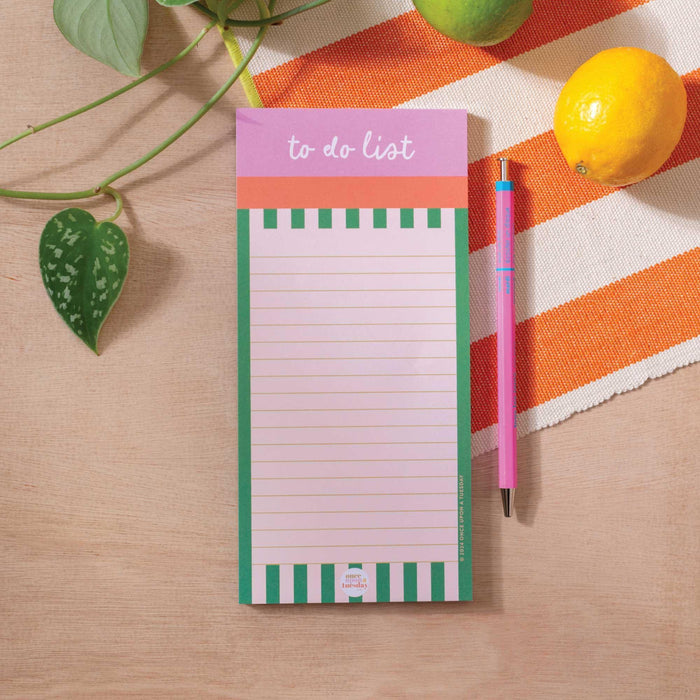 A Cabana Stripe To Do List Pad with green and white stripes and pink header, displayed on a wooden desk surface with an orange striped textile, pink pen, lemon, and potted plant leaf in view