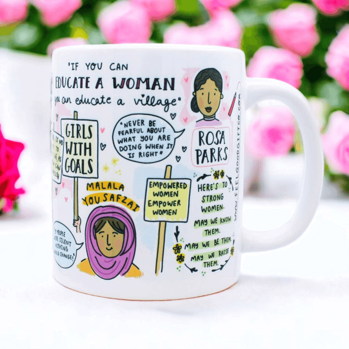 White ceramic mug featuring inspiring quotes about women's education and empowerment, decorated with hand-drawn illustrations and protest-style signs. Displayed against vibrant pink roses and green foliage background. The mug shows hand-lettered text and whimsical sketches in a modern doodle style.