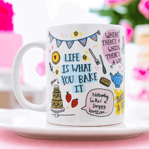 White ceramic mug with playful baking illustrations including British bunting, a tiered cake, strawberries, and teapot, featuring the quote "Life is what you bake it" and other baking puns, displayed on a pink surface with soft floral background.