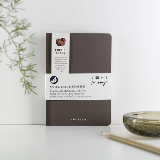 A minimalist brown A5 notebook made from recycled coffee beans and paper, featuring the SUCSEED logo in silver. The notebook is displayed on a white surface with a decorative green plant and gold pen in the background. The packaging band shows VENT's logo and 'Notes, Lists & Scribbles' text.