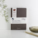 A minimalist brown A5 notebook made from recycled coffee beans and paper, featuring the SUCSEED logo in silver. The notebook is displayed on a white surface with a decorative green plant and gold pen in the background. The packaging band shows VENT's logo and 'Notes, Lists & Scribbles' text.